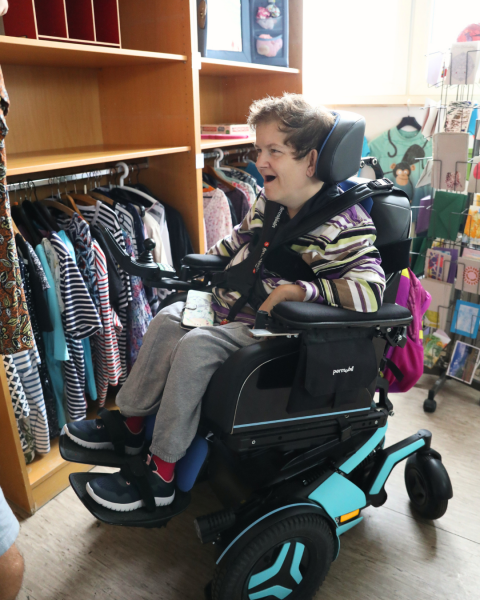 Eine Person im Rollstuhl begutachtet lächelnd ein Oberteil aus der Second-Hand-Klamotten-Abteilung.