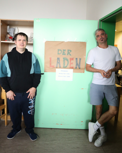 Zwei Personen stehen neben der Eingangstür vom Laden und schauen in die Kamera. Auf der Eingangstür ist ein großes Plakat. Darauf steht in bunten Buchstaben: „Der Laden“. Darunter ist ein kleineres Plakat. Darauf steht: „Öffnungszeiten. Montag 11 bis 12 Uhr. Mittwoch 11 bis 12 Uhr.“