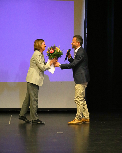 Zwei Personen stehen auf einer Bühne. Eine Person überreicht der anderen gerade einen Blumenstrauß. Beide lächeln sich gegenseitig an.