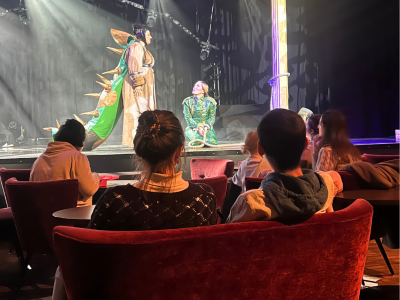 Auf der Bühne im GOP kniet ein kleiner Drache vor einem großen Drachen. Im Publikum sitzen Kinder und Erwachsene auf Sesseln und Sofas, die dem Schauspiel zusehen.