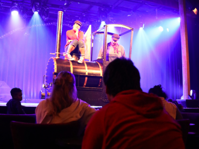 Lukas, der Lokomotivführer, sitzt in seiner Lok Emma. Jim Knopf sitzt vorne auf der Lok. Beide und Emma sind auf der Bühne. Im Publikum sitzen Menschen, die zugucken.