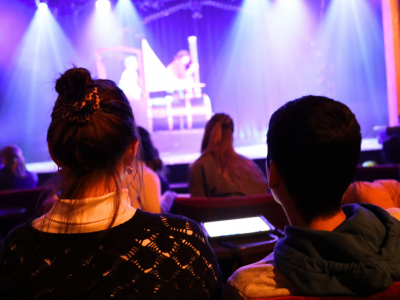 Von hinten fotografiert sitzt eine Person neben einem Kind auf einem Sofa. Hinten ist unscharf die Bühne mit Lukas, dem Lokomotivführer, Jim Knopf und der Lok Emma.