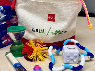 Ein Stimming-Kit mit Logi vom GOP sowie dem Doppellogo von der GiB und dem AZH steht auf einem Tisch. Davor liegen viele Materialien wie Fidget Toys, die autistischen Menschen bei der Stimulation helfen.